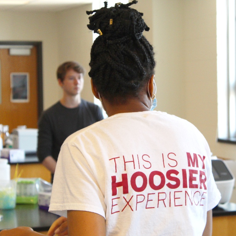 Female student facing away wearing a t-shirt that says This is My Hoosier Experience. A blurred male student stands in the background.