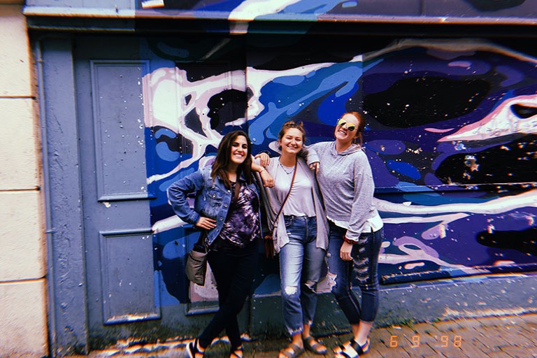 Katie Rodwell and two coworkers posing for a photo in front of a wall art piece on the side of a building